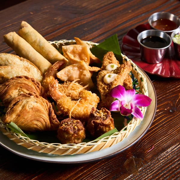A woven basket of assorted Thai appetizers, including spring rolls, curry puffs, and fried shrimp, with dipping sauces.