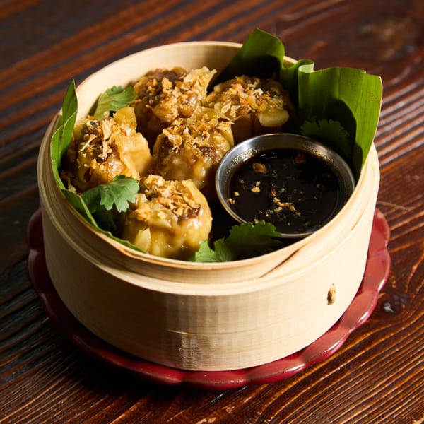 Steamed Thai dumplings with crispy garlic and cilantro, served with a dark dipping sauce in a bamboo steamer.