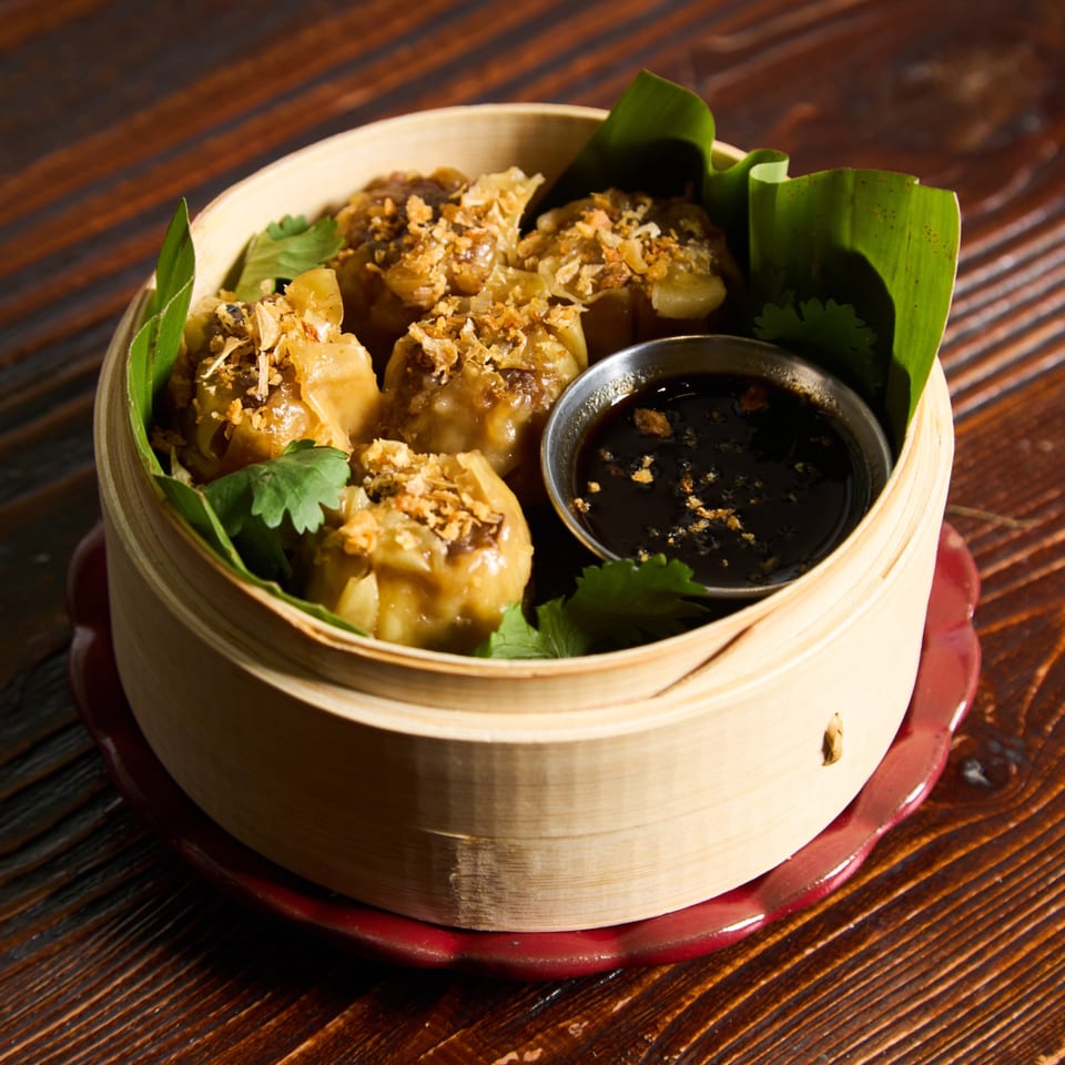 Steamed Thai dumplings with crispy garlic and cilantro, served with a dark dipping sauce in a bamboo steamer.