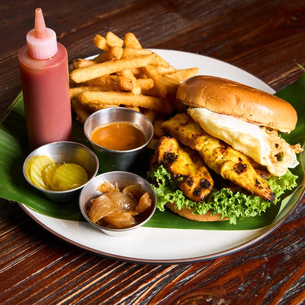 Grilled chicken sandwich with lettuce, fried egg, fries, and dipping sauces on a banana leaf.