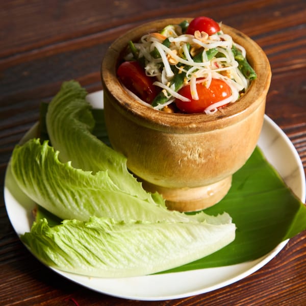 Som Tum (papaya salad) in a wooden mortar, served with fresh lettuce on a banana leaf.