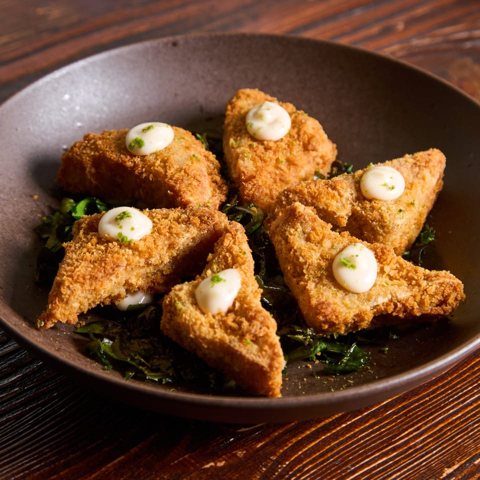 Crispy golden-brown fried tofu triangles with creamy white sauce and green garnish, served on dark greens.